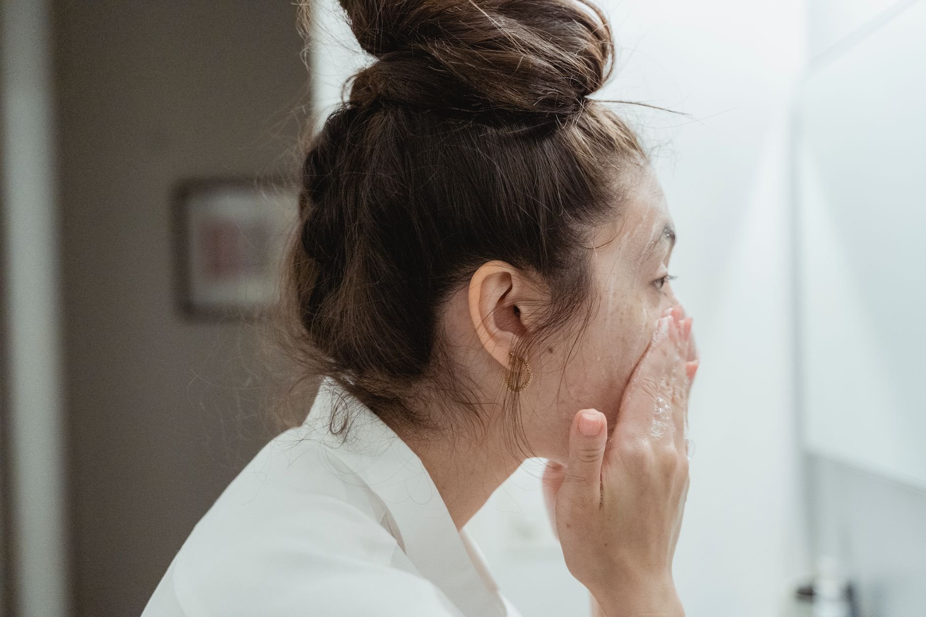A young woman in a bathrobe follows her skincare routine, washing her face in a sunlit bathroom
